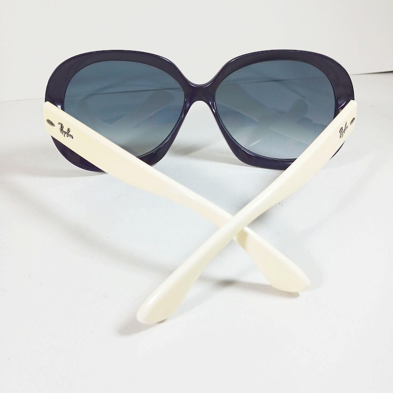 Peut inclure: Lunettes de soleil surdimensionn&eacute;es avec une monture violet fonc&eacute; et des verres d&eacute;grad&eacute;s. Les branches sont blanc cass&eacute; et se croisent au centre. Le logo Ray-Ban est visible sur les branches.