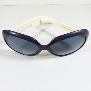 Peut inclure: Une paire de lunettes de soleil avec une monture violet fonc&eacute; et des verres gris. Les branches sont blanc cass&eacute;. Les lunettes de soleil sont pos&eacute;es sur un fond blanc. Les lunettes de soleil sont de style &oelig;il de chat.