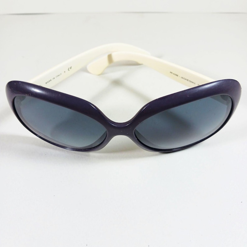 Peut inclure: Une paire de lunettes de soleil avec une monture violet fonc&eacute; et des verres gris. Les branches sont blanc cass&eacute;. Les lunettes de soleil sont pos&eacute;es sur un fond blanc. Les lunettes de soleil sont de style &oelig;il de chat.