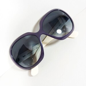 Peut inclure: Lunettes de soleil violettes avec des verres fonc&eacute;s et des branches blanc cass&eacute;. Les lunettes ont une monture arrondie et surdimensionn&eacute;e. Le texte "MADE IN ITALY" et le logo Ray-Ban sont visibles &agrave; l'int&eacute;rieur de la monture.