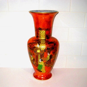 Puede incluir: Un jarrón de cerámica naranja con la parte superior acampanada y base redonda. El jarrón presenta una ilustración detallada de figuras con vestimenta tradicional. Los detalles dorados y el borde azul añaden atractivo decorativo a esta pieza.