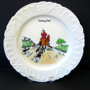 Puede incluir: Un plato de cerámica blanco con un borde elevado y un diseño decorativo. El plato presenta una ilustración colorida de un grupo de personas a caballo con perros, y el texto "Setting Out."