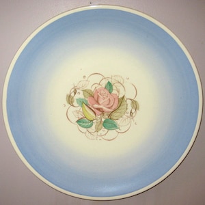 Pode incluir: Um prato de cerâmica azul e branco com uma rosa rosa e folhas verdes no centro. O prato tem uma borda decorativa de volutas e folhas marrons.