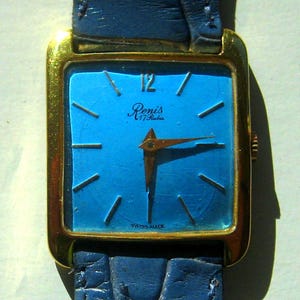 Puede incluir: Un reloj cuadrado dorado con una esfera azul y agujas doradas. La esfera del reloj tiene el texto "Renis 17 Rubis" y "Swiss Made".