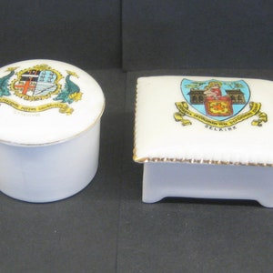 May include: Two white ceramic trinket boxes. One is round with a lid, the other is square. Both have gold trim and feature colorful crests with text. The round box has a crest with dolphins and the text "SALUBRITATE". The square box has a crest with a building and the text "SELKIRK".