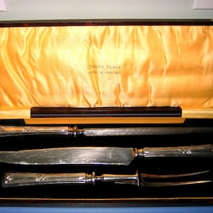 May include: A vintage carving set in a wooden case. The set includes a carving knife, a sharpening steel, and a carving fork. The case is lined with gold fabric and has the text "COUNTY PLATE MADE IN ENGLAND".