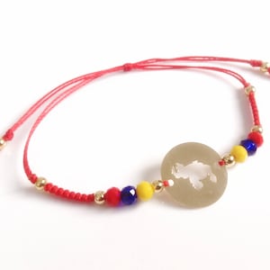 May include: Red string bracelet with a gold charm featuring a cut-out map design. The bracelet is accented with yellow, red, and blue beads.