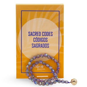 May include: A beaded bracelet with iridescent, faceted beads in shades of purple and blue, next to a yellow card. The card has the text "SACRED CODES CÓDIGOS SAGRADOS" in blue. The bracelet has a gold-colored bead at the end.