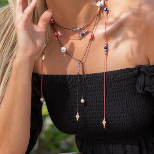 May include: Layered necklace with red and blue evil eye beads and gold hamsa charms. The necklace is worn by a person with long blonde hair.