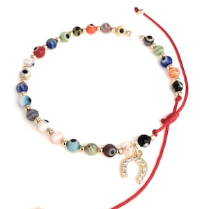 May include: A colorful beaded bracelet featuring various eye-shaped beads in shades of blue, green, red, and white. The bracelet includes gold-colored beads and a charm of a horseshoe embellished with small, clear stones. The bracelet is strung on a red cord.