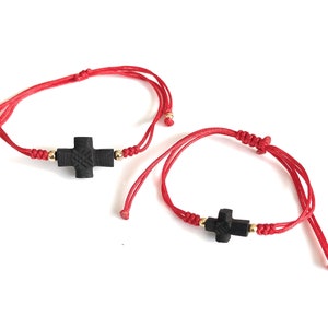 May include: Two red string bracelets with black wooden cross charms. The bracelets are adjustable and have gold beads.
