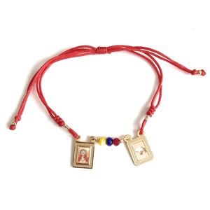 Puede incluir: Pulsera de cuerda roja con dijes de oro. Un dije presenta una imagen religiosa y el otro dije presenta un recorte de un país. La pulsera tiene cuentas amarillas, azules y rojas.