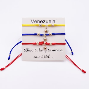May include: Three colorful string bracelets with gold star charms and beads. The bracelets are yellow, blue, and red, representing the colors of the Venezuelan flag. A card with the word "Venezuela" and the phrase "Llevo tu luz y tu aroma en mi piel..." is displayed behind the bracelets.