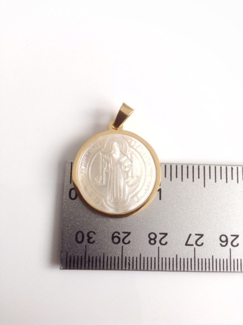 May include: A gold-toned pendant with a white, round, mother-of-pearl inlay featuring a religious figure. The pendant is approximately 2.8 cm in diameter.