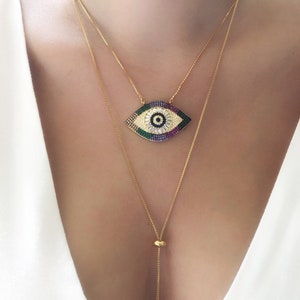 May include: A gold chain necklace with a large, colorful evil eye pendant. The pendant is made of rhinestones and has a blue center. The necklace is layered with a second, thinner gold chain.