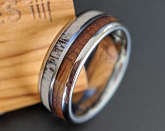 May include: A silver and brown ring with a wood inlay and a white antler inlay. The ring is on a wooden surface.
