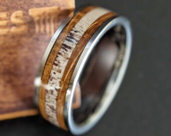May include: A close-up of a wooden and stainless steel ring. The ring has a brown wood inlay with white accents. The ring is on a wooden surface.