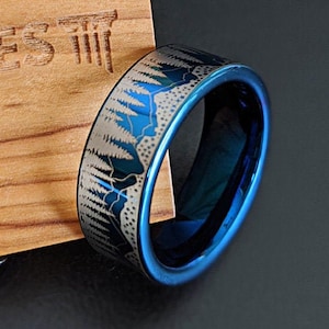 May include: A blue and silver tungsten carbide ring with a forest scene design. The ring is on a wooden surface.