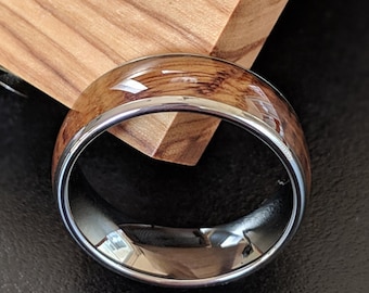 May include: A ring featuring a polished dark metallic band with a smooth wood grain inlay. The ring is positioned against a light wood block. The text PILLAR STYLES is printed in white at the bottom of the image with a pillar icon on each side.