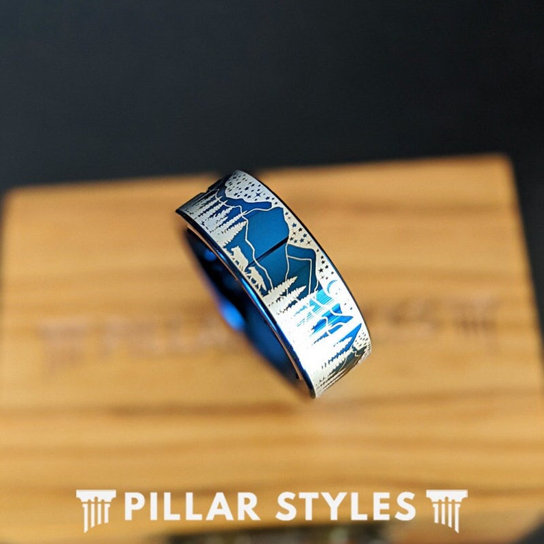 May include: A blue tungsten carbide ring with a silver mountain and forest design. The ring is engraved with a moon and stars.
