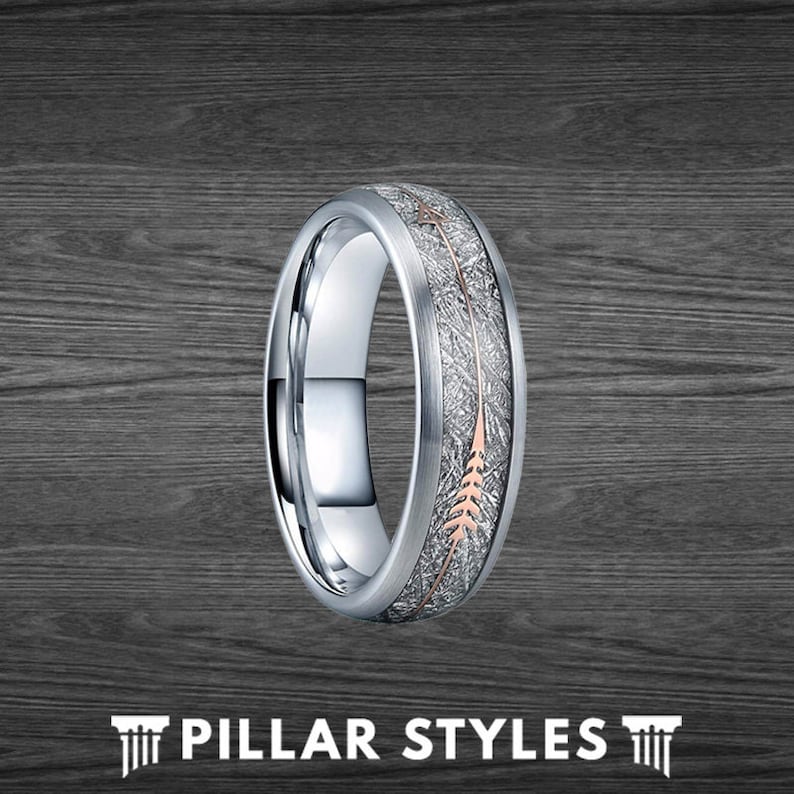 May include: A silver wedding band with a rose gold arrow design. The band has a textured finish.