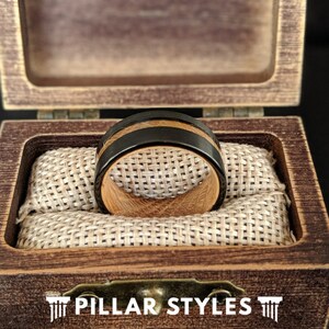 May include: A wooden ring with a black inlay, displayed in a wooden box with a brown fabric lining. The box has the text "PILLAR STYLES" on the front.