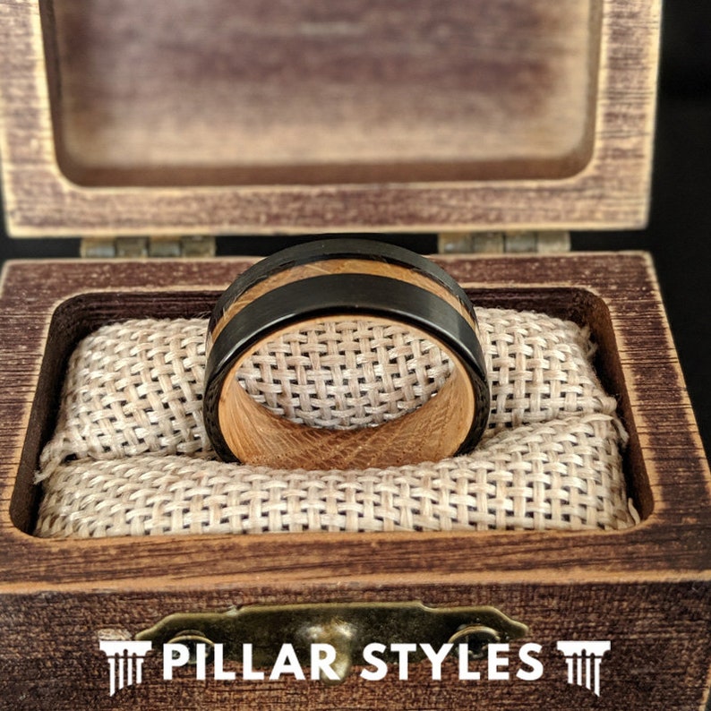 May include: A wooden ring with a black inlay, displayed in a wooden box with a brown fabric lining. The box has the text "PILLAR STYLES" on the front.