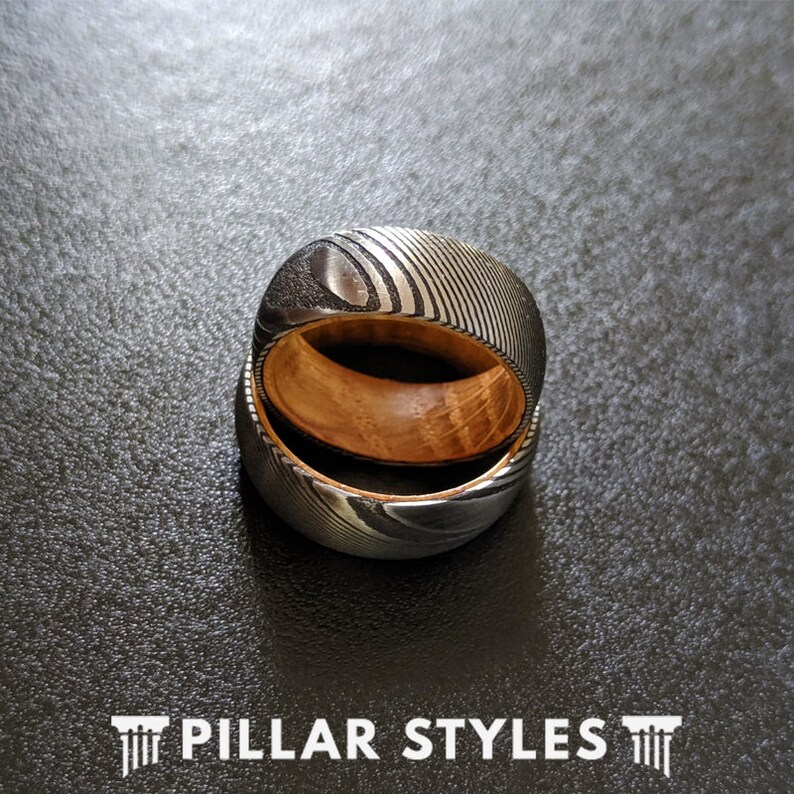 May include: Two stacked rings with a wood inlay and a patterned metal exterior. The rings are silver and black with a wood grain pattern.