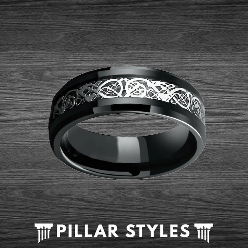 May include: A black tungsten carbide ring with a silver Celtic knot design. The ring is polished and has a beveled edge.