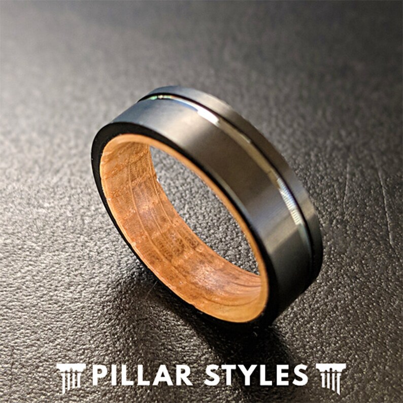 May include: A black and wood ring with a silver band. The ring is made of black tungsten carbide and has a wood inlay. The wood inlay is a light brown color and has a natural grain pattern.