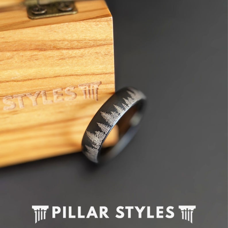 May include: A black ring with a forest design etched around the band. The ring is displayed next to a wooden box. The text "STYLES" is visible on the box. The text "PILLAR STYLES" is at the bottom of the image.