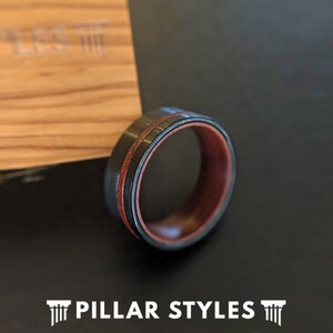 May include: A black and red wood ring with a black metal band. The ring is made by Pillar Styles.