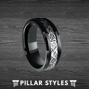 May include: A black tungsten carbide ring with a silver Celtic knot design. The ring is polished and has a beveled edge.