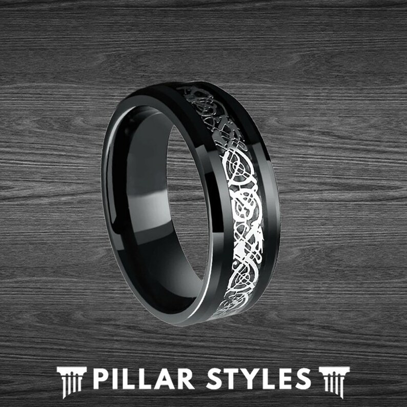 May include: A black tungsten carbide ring with a silver Celtic knot design. The ring is polished and has a beveled edge.