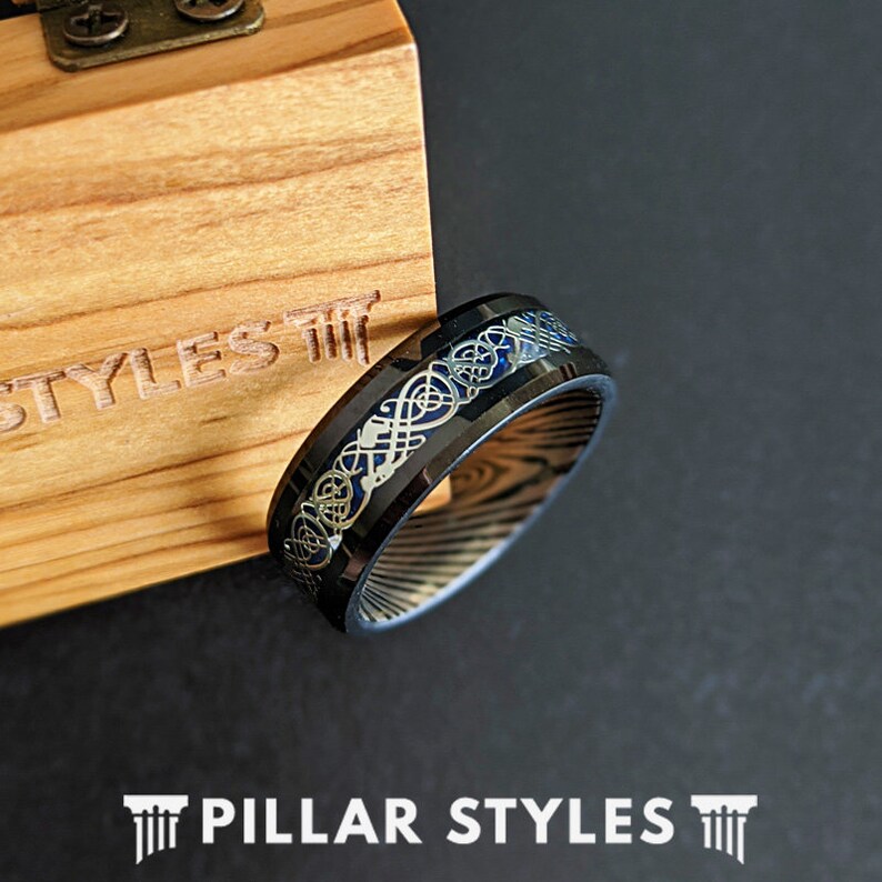 May include: A black and blue tungsten carbide ring with a silver Celtic knot design. The ring is shown on a wooden box with the text "Pillar Styles" on the front.