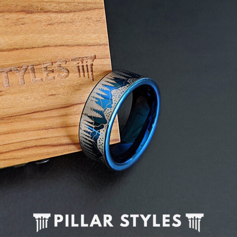 May include: A blue and silver tungsten carbide ring with a forest scene design. The ring is on a wooden surface.