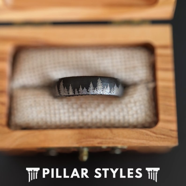 May include: A black ring with a forest design in a wooden box. The ring features a band with a detailed white tree line pattern. The ring is displayed on a beige fabric inside a light brown wooden box. The text "PILLAR STYLES" is at the bottom.