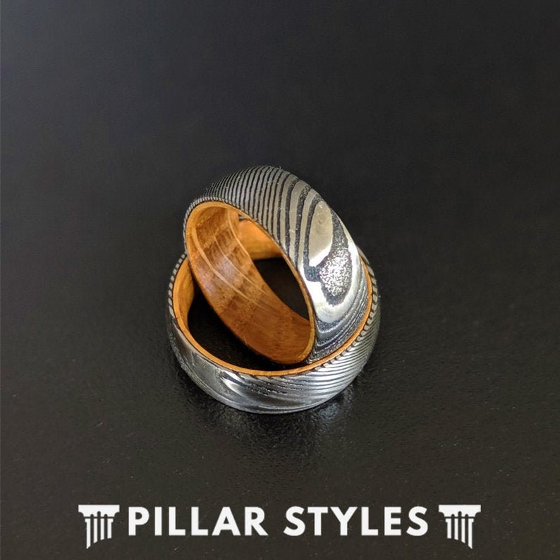 May include: Two stacked rings, one with a wood inlay and the other with a patterned metal finish. The rings are a dark grey color with a silver sheen.