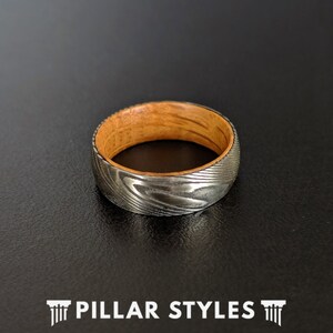 May include: A silver and wood ring with a patterned design. The ring is made of Damascus steel and wood.