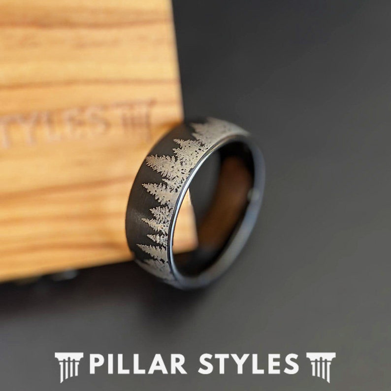 May include: A black ring with a forest design etched around the band. The ring is set against a wooden background and a dark gray surface. The text "PILLAR STYLES" is at the bottom of the image.