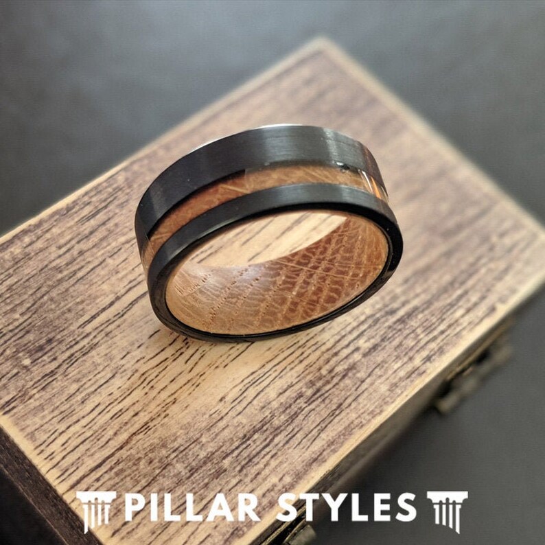 May include: A black and brown wooden ring with a black metal band. The ring is sitting on a wooden box with the text "PILLAR STYLES" on it.