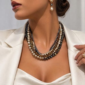May include: A multi-strand pearl necklace featuring layers of cream, bronze, and dark grey pearls. The set includes a matching drop earring with a gold-tone post and a pearl, along with a gold-tone ring featuring a cluster of small white pearls.
