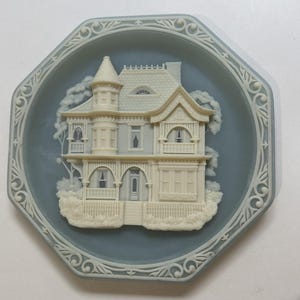 May include: A decorative, octagonal plate with a bas-relief of a Victorian house. The house is off-white against a light blue background, with intricate details. The plate's border features a white, ornate design.
