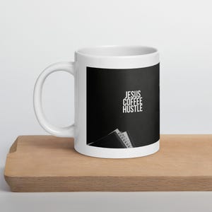 May include: White ceramic coffee mug with the words "JESUS COFFEE HUSTLE" in white text on a black background. The mug is sitting on a wooden surface.