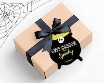 May include: A beige gift box with a black ribbon and bow. A black tag with a cauldron design and the words "Witching you a Spooky Halloween" is attached to the ribbon. A spiderweb illustration is in the background.
