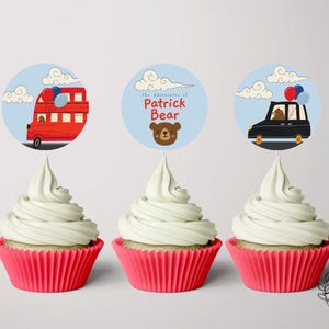 Puede incluir: Tres cupcakes con forros rojos y glaseado blanco. Cada cupcake tiene un adorno de papel redondo y colorido con un diseño diferente. Un adorno presenta un autobús de dos pisos rojo, otro un taxi negro y el tercero un oso marrón con el texto "The Adventures of Patrick Bear".