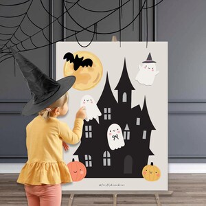 Puede incluir: Un póster de Halloween con una casa embrujada negra, una luna llena, un murciélago y fantasmas amigables. Dos calabazas naranjas están en la parte inferior. Una niña con un sombrero de bruja interactúa con el póster.