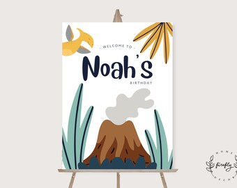May include: A welcome sign for a dinosaur themed birthday party. The sign is white with a brown volcano, green plants, and a yellow dinosaur. The text on the sign reads "Welcome to Noah's Birthday".