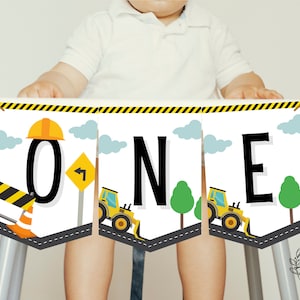 Puede incluir: Pancarta de primer cumpleaños con temática de construcción con la palabra "ONE" en letras negras. La pancarta presenta vehículos de construcción amarillos, un casco y una señal de tráfico.