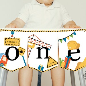 Puede incluir: Una pancarta con temática de construcción para una fiesta de primer cumpleaños. La pancarta está hecha de cartulina blanca con rayas amarillas y negras. Las letras "one" son negras y están rodeadas de imágenes con temática de construcción, incluyendo una grúa, un camión y un cono de construcción. La pancarta también incluye las palabras "caution" y un casco amarillo.